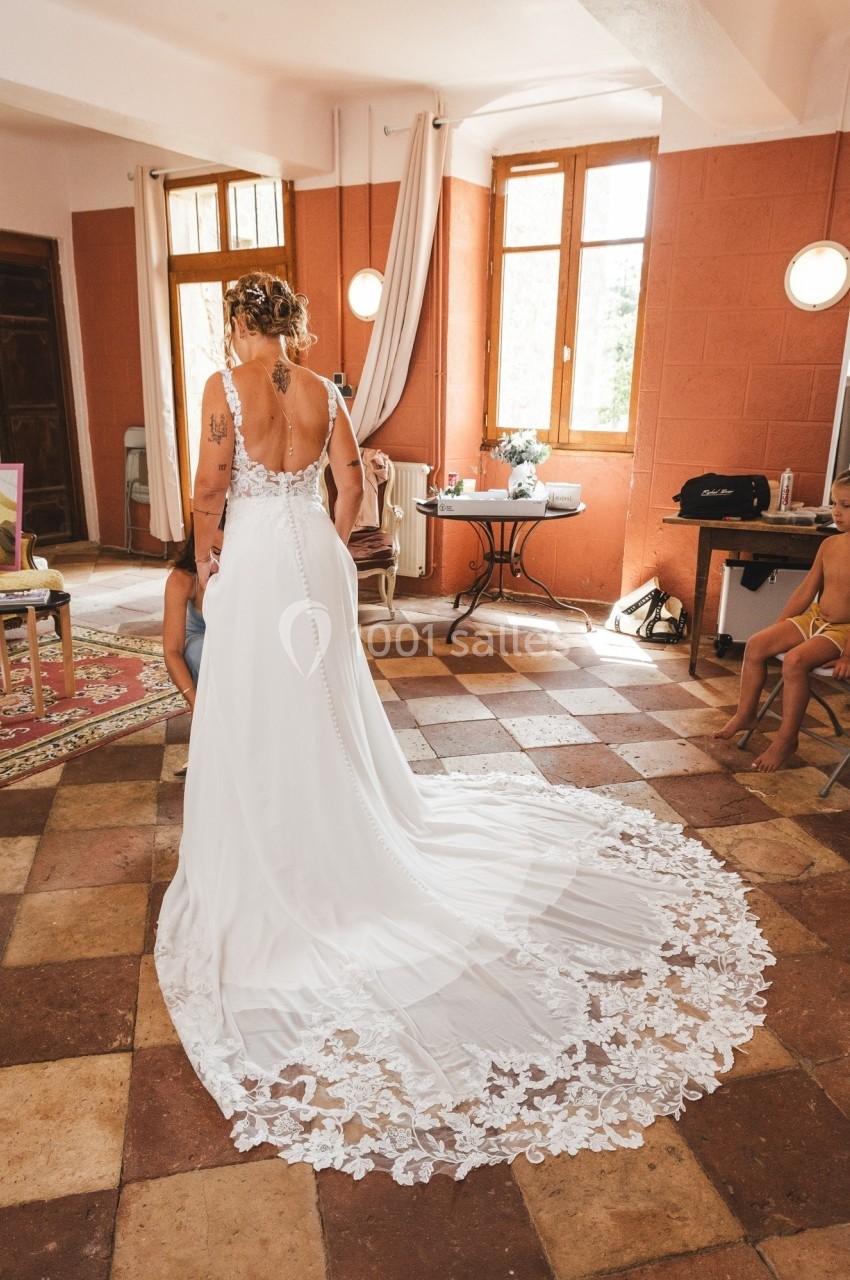 Une femme en robe de mariée blanche avec une longue traîne en dentelle, vue de dos dans une pièce lumineuse.