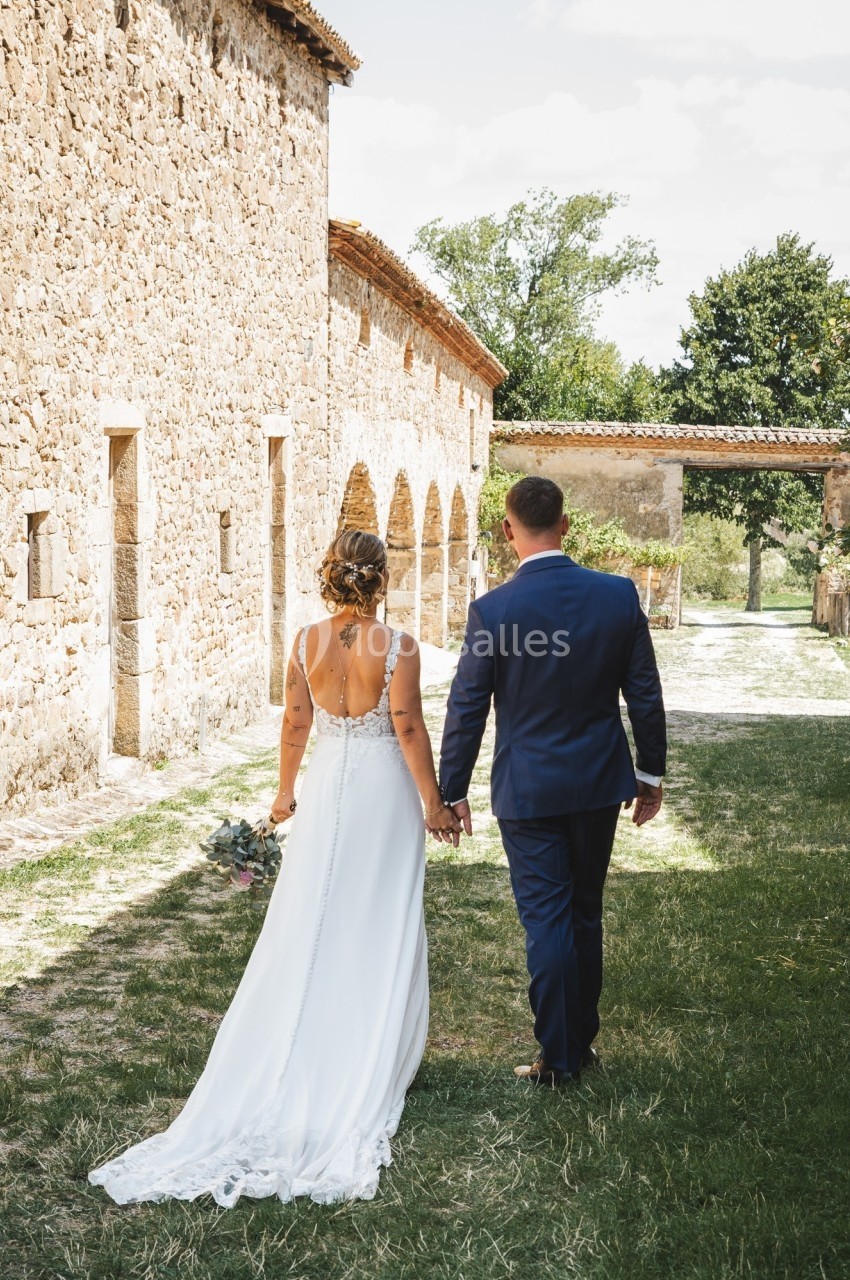 Un couple en tenue de mariage marche main dans la main dans une cour bordée de bâtiments en pierre.