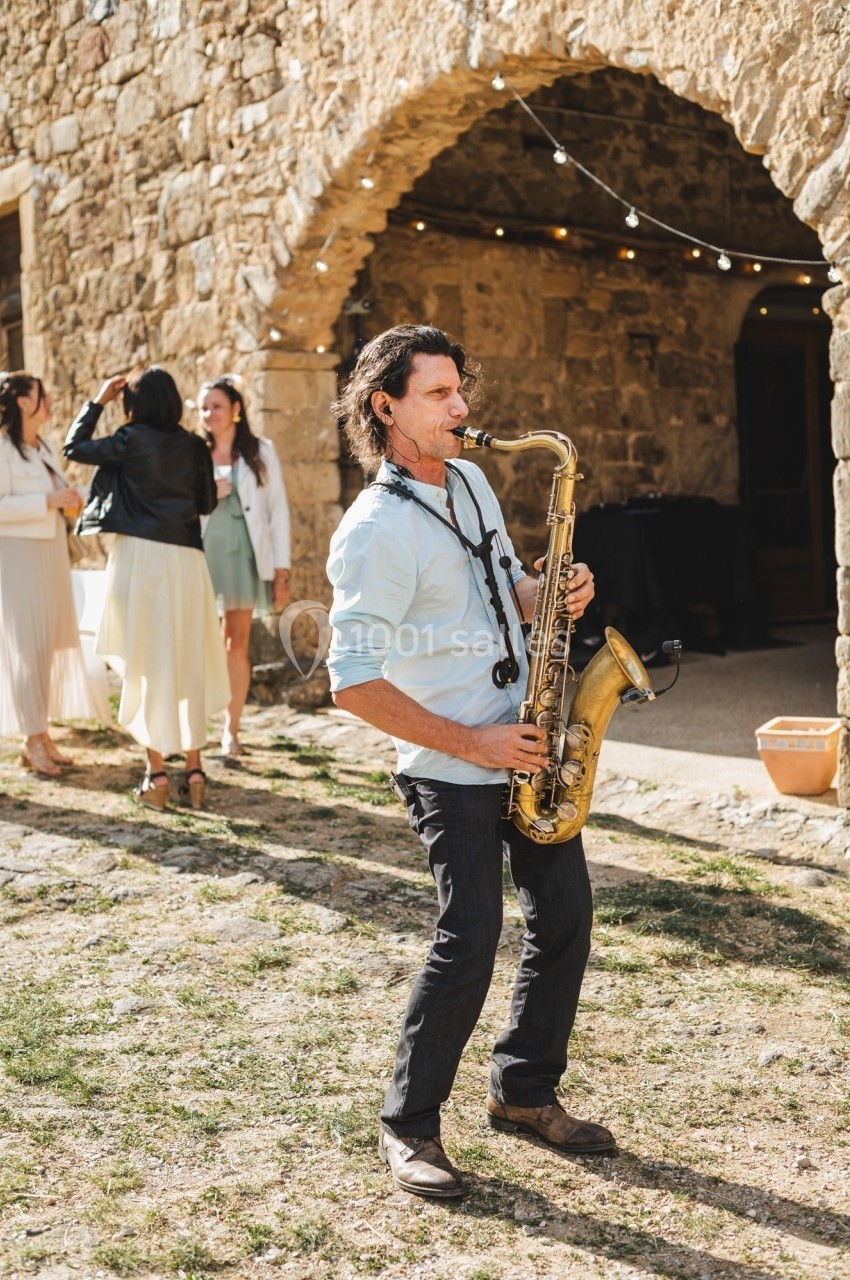 Un musicien joue du saxophone devant un bâtiment en pierre, avec des personnes discutant en arrière-plan.