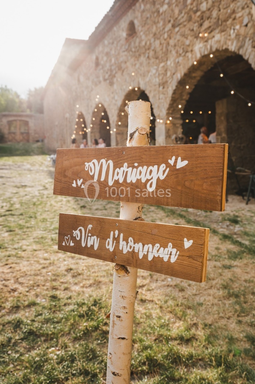 Panneaux en bois indiquant ’Mariage’ et ’Vin d'honneur’ devant un bâtiment en pierre avec guirlandes lumineuses.