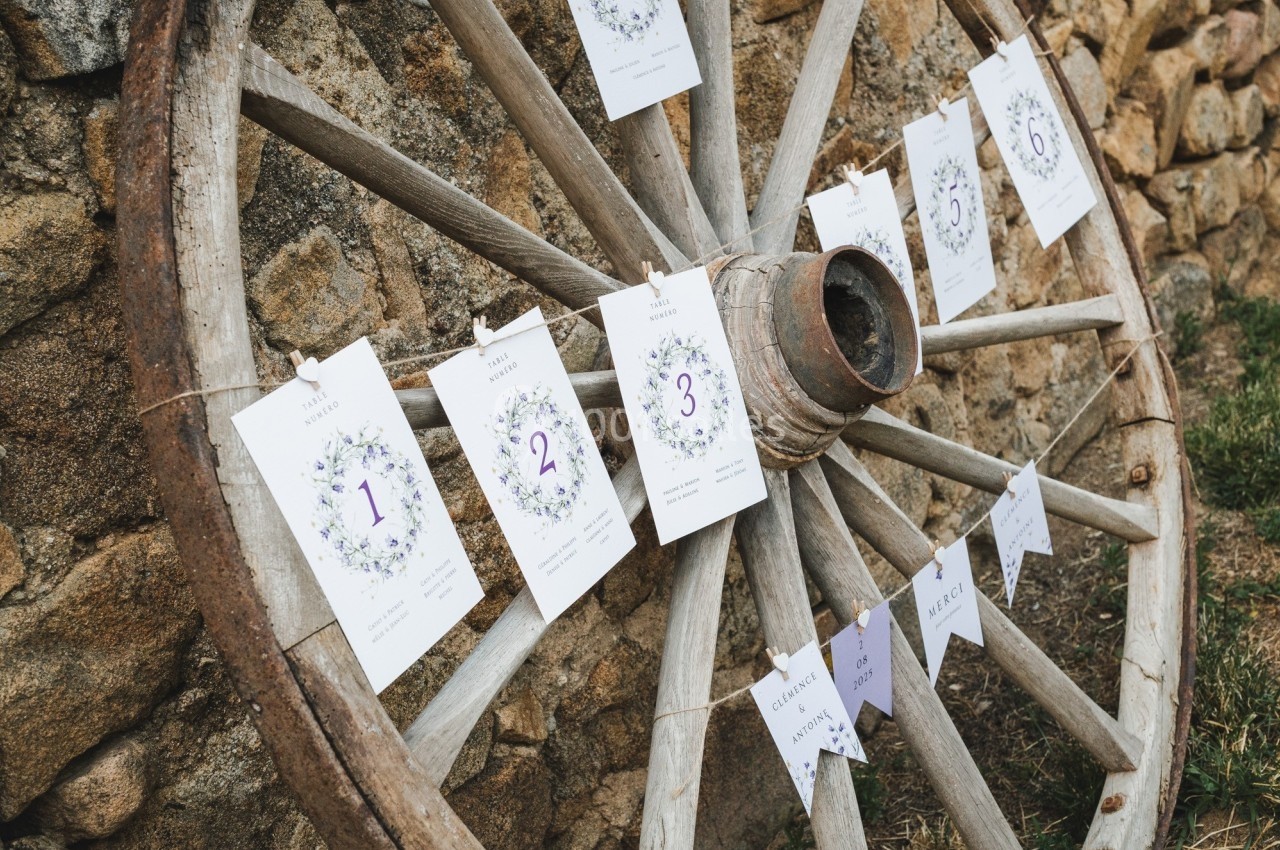 Vieille roue en bois décorée de cartes numérotées et de fanions, posée contre un mur en pierre.