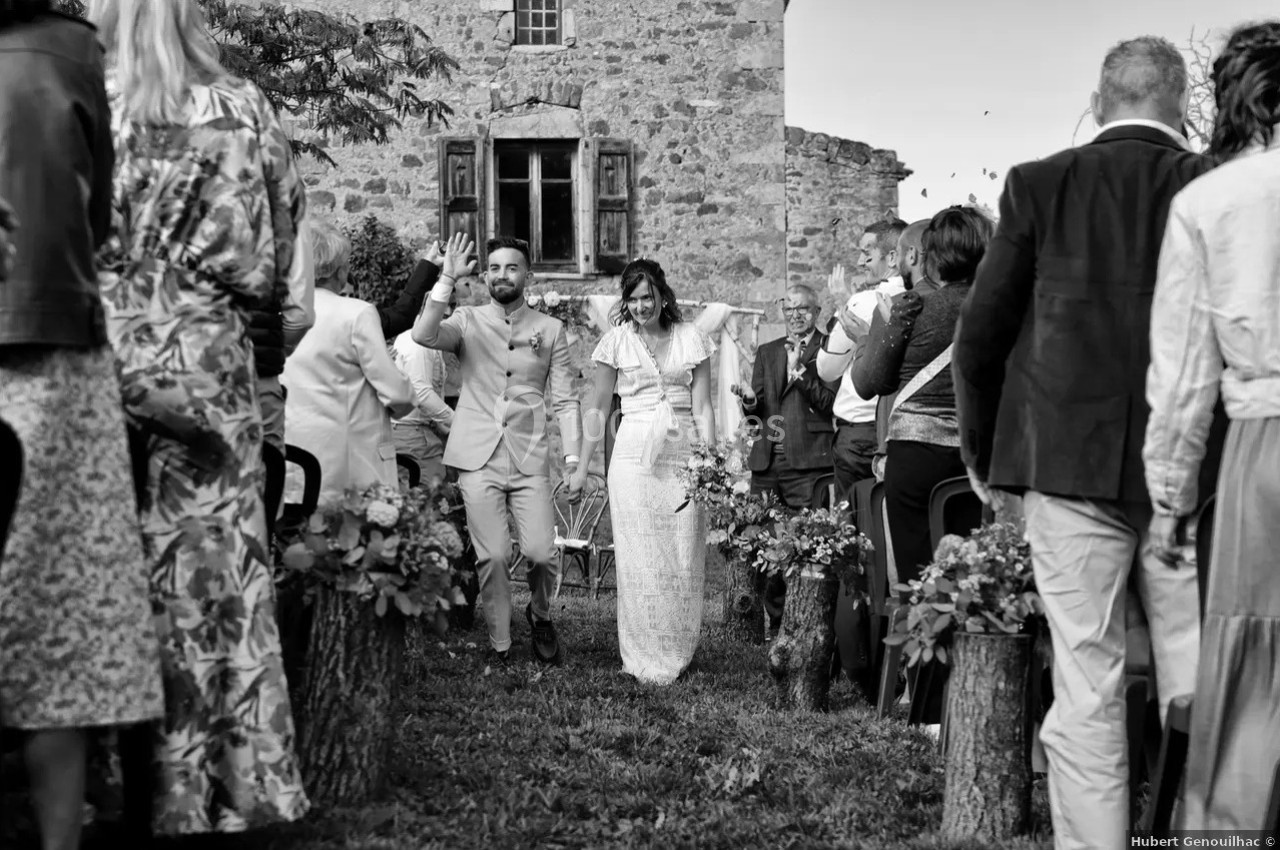 Un couple souriant marche entre des invités lors d'une cérémonie en extérieur devant un bâtiment en pierre.