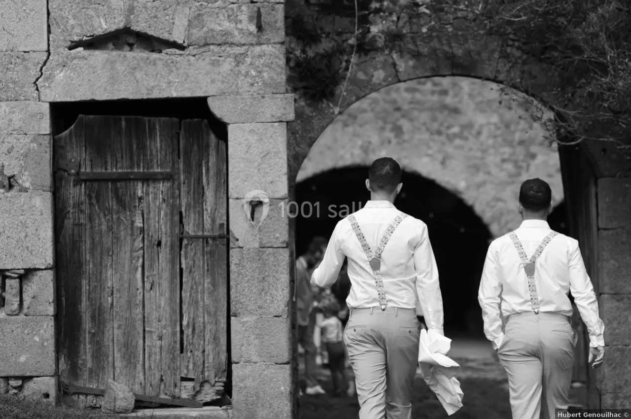 Deux hommes en chemises blanches et bretelles marchent sous une arche en pierre dans un cadre rustique.