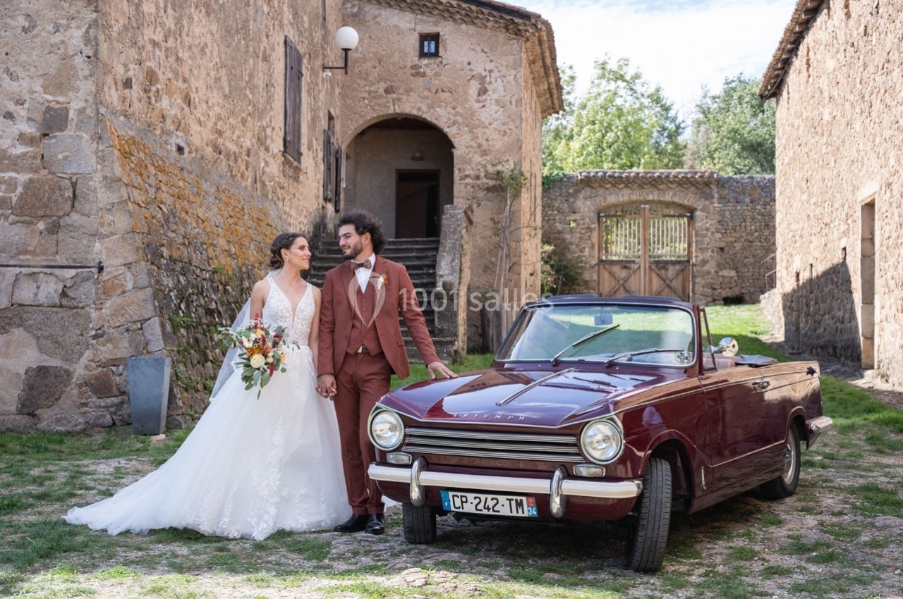 Un couple de mariés pose près d'une voiture ancienne rouge dans une cour en pierre.