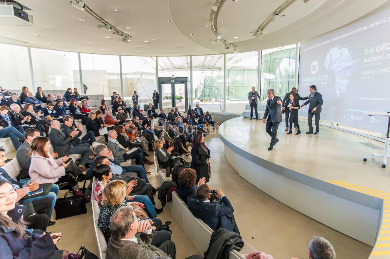 Personnes assistant à une conférence dans un amphithéâtre moderne, avec un intervenant debout sur scène.