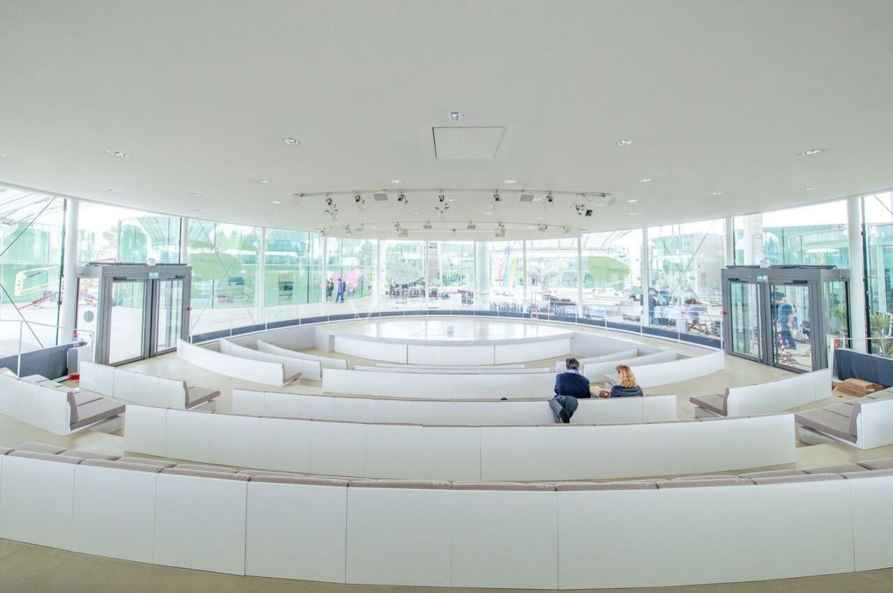 Salle circulaire lumineuse avec des sièges blancs disposés en gradins, entourée de grandes baies vitrées.