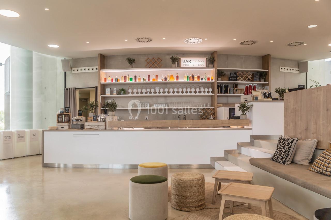 Intérieur d'un café moderne avec comptoir blanc, étagères garnies de bouteilles et espace assis avec coussins.