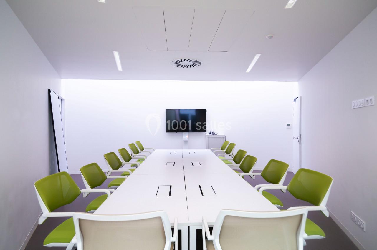 Salle de réunion moderne avec une grande table blanche, des chaises vertes et un écran mural.