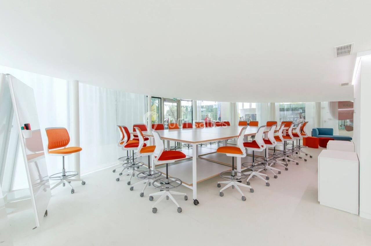 Salle de réunion lumineuse avec une grande table blanche entourée de chaises orange et blanches sur roulettes.