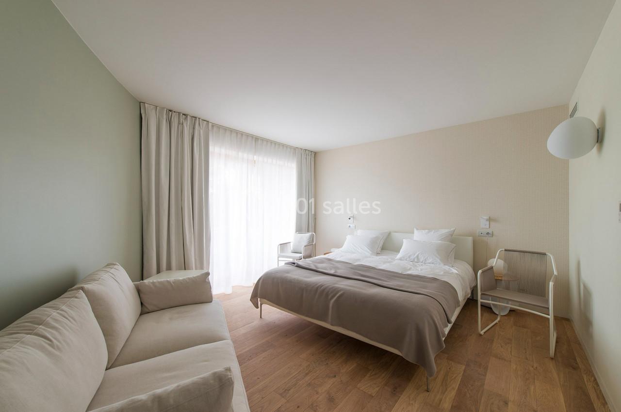 Chambre lumineuse avec un lit double, un canapé beige, des rideaux clairs et un sol en parquet.