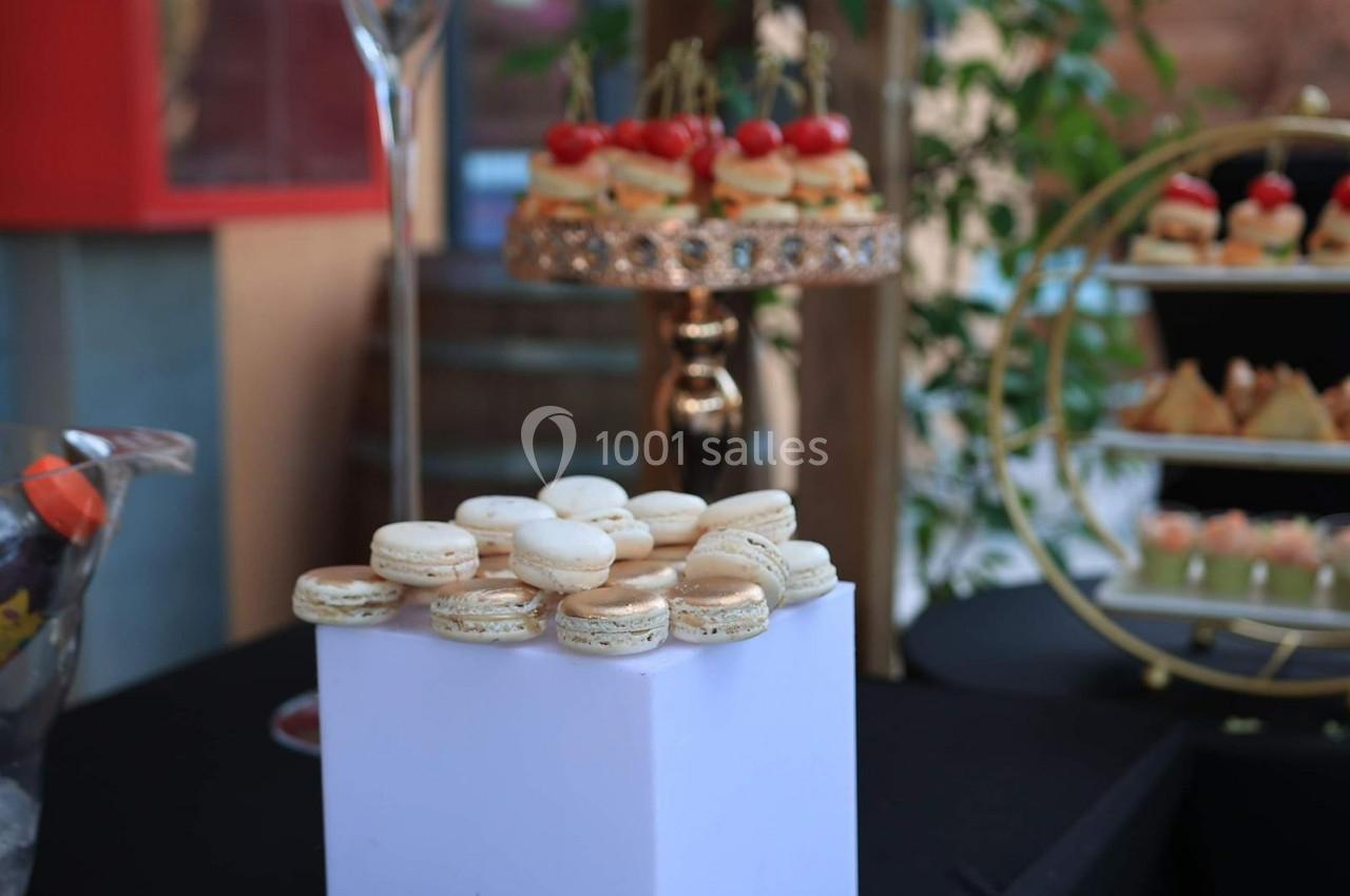 Plateau de macarons blancs et dorés disposés sur un socle blanc, avec des amuse-bouches en arrière-plan.