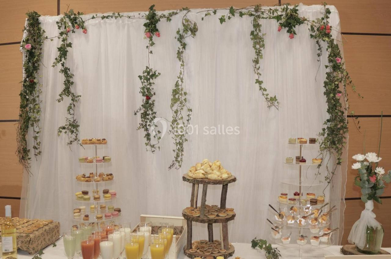 Buffet décoré avec rideau blanc, guirlandes de feuillage, verrines, jus de fruits et pâtisseries sur des présentoirs.
