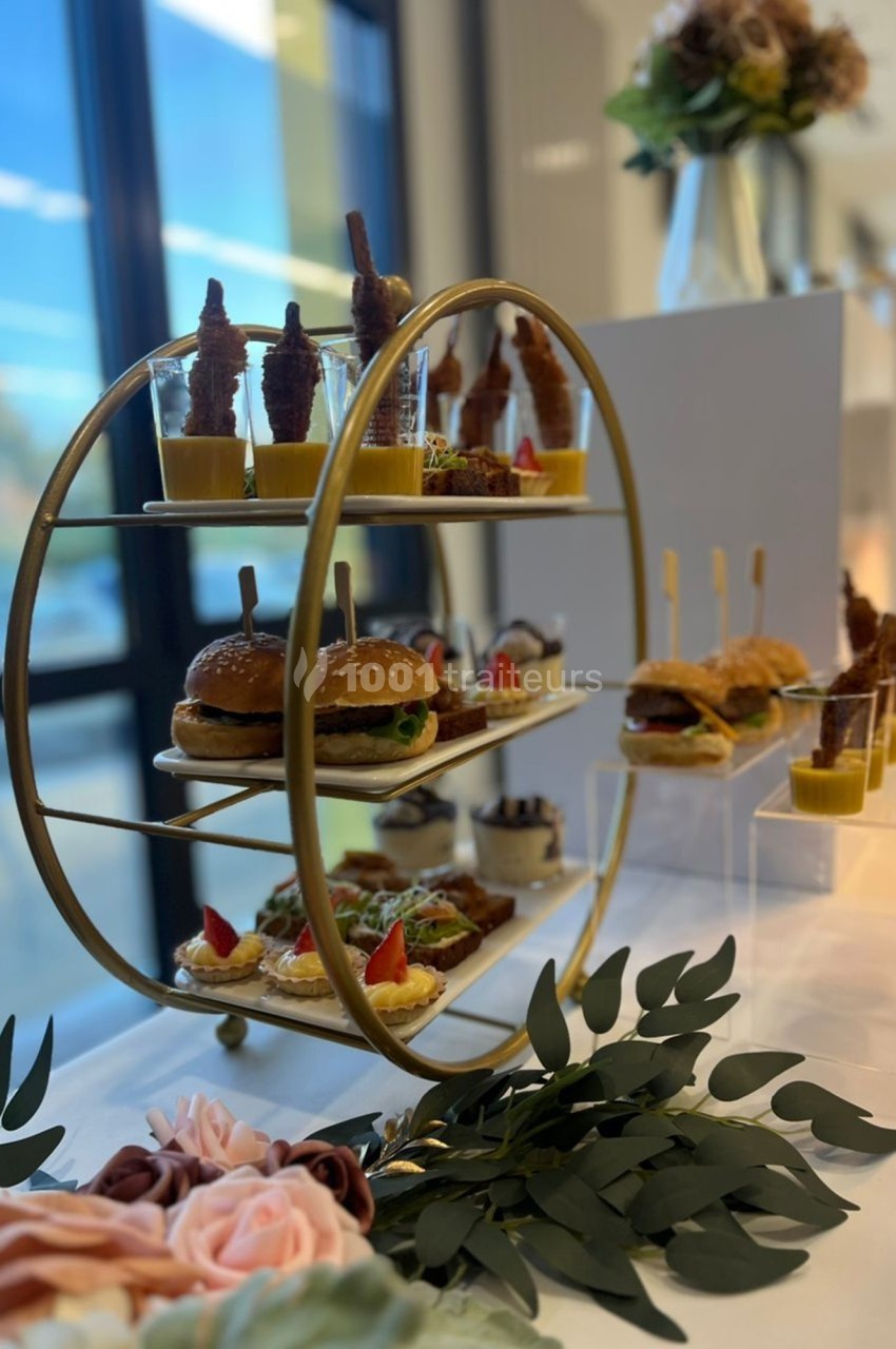 Présentoir doré avec des amuse-bouches variés, incluant mini-burgers, verrines et brochettes, sur une table décorée.