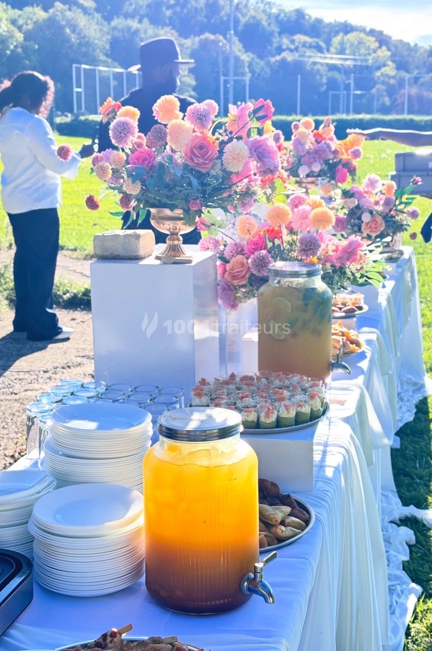 Table décorée en extérieur avec des fleurs colorées, des boissons en jarres et des assiettes empilées pour un buffet.