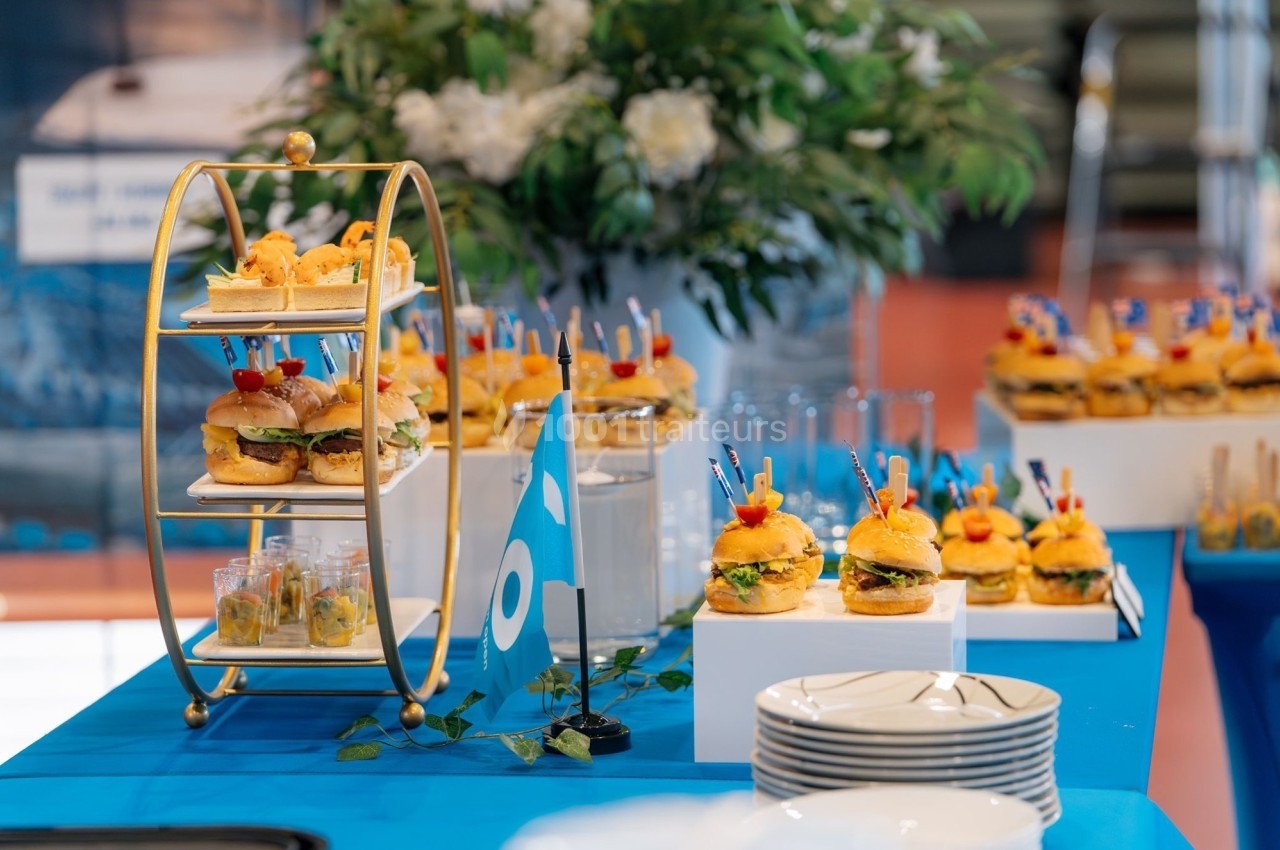 Plateau de mini-burgers et verrines sur une table décorée en bleu, avec des fleurs en arrière-plan.