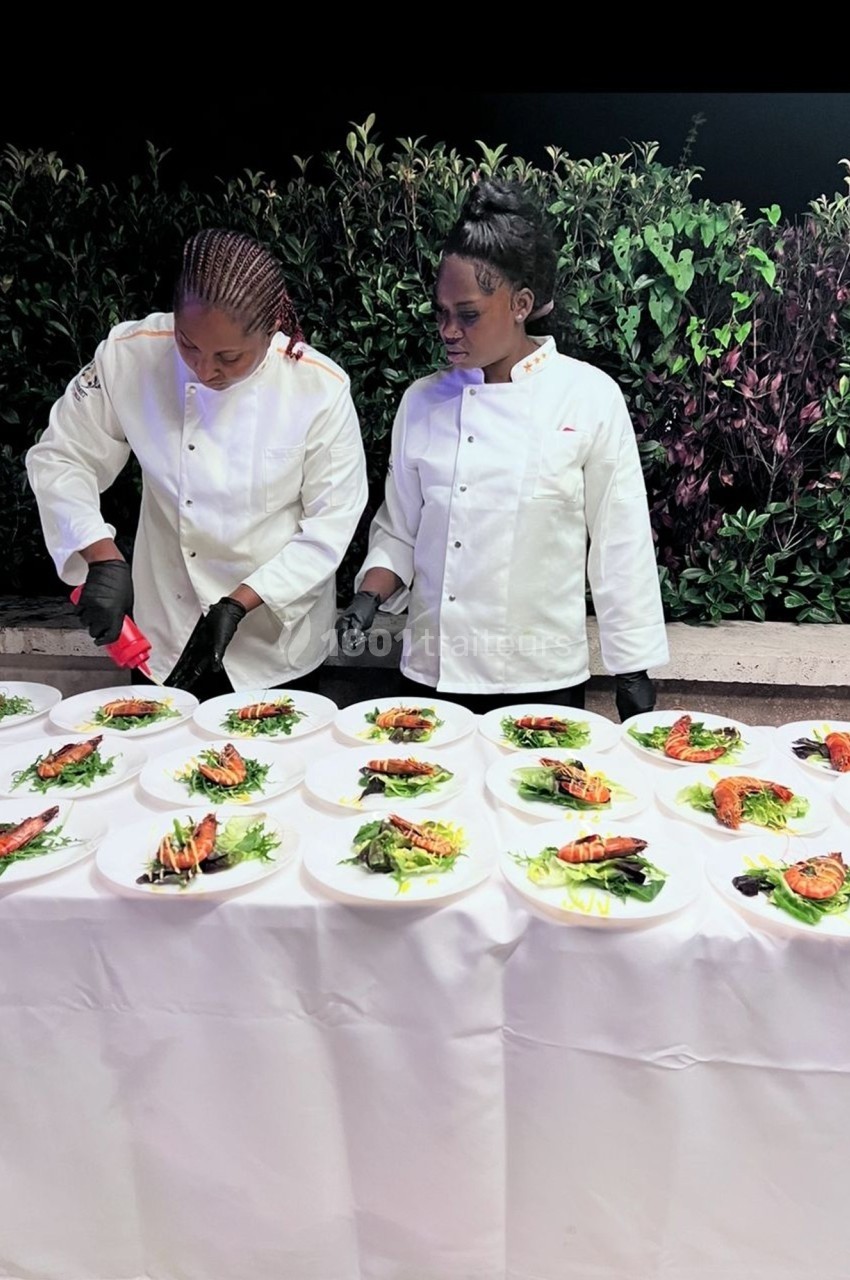 Deux chefs en tenue blanche dressent des assiettes élégantes sur une longue table, en extérieur, devant des plantes.
