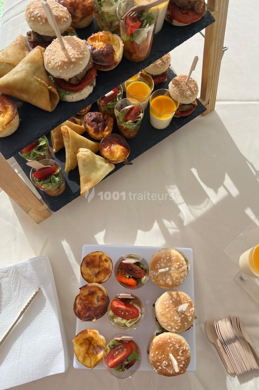 Plateau de mini-burgers, samoussas, quiches et verrines sucrées et salées sur une table ensoleillée.