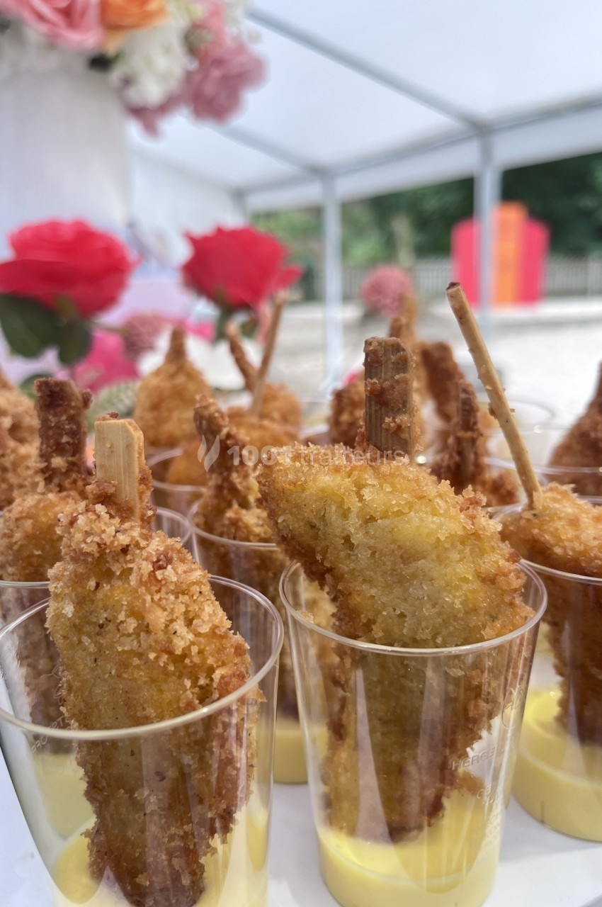 Bouchées de poulet pané sur bâtonnets, servies dans des verrines avec une sauce jaune, décor floral en arrière-plan.