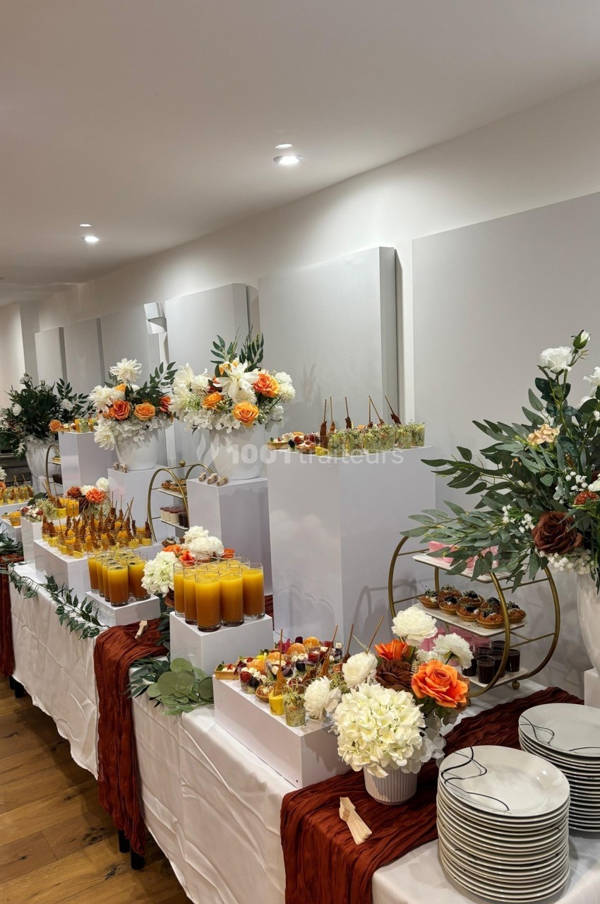 Table de buffet élégante avec fleurs, verrines, jus d'orange et desserts variés, décorée dans des tons chauds et naturels.