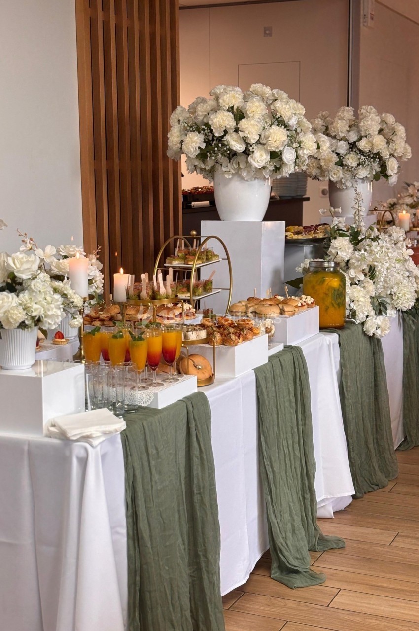 Table décorée avec des fleurs blanches, nappes blanches et chemins de table verts, garnie de boissons et mets variés.