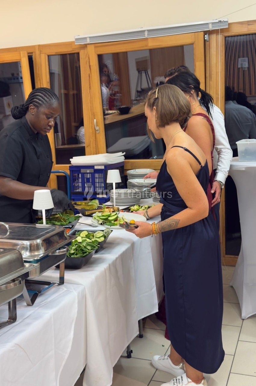 Une cuisinière sert des plats à des convives dans un buffet, avec des salades et des plats chauds disposés sur une table.