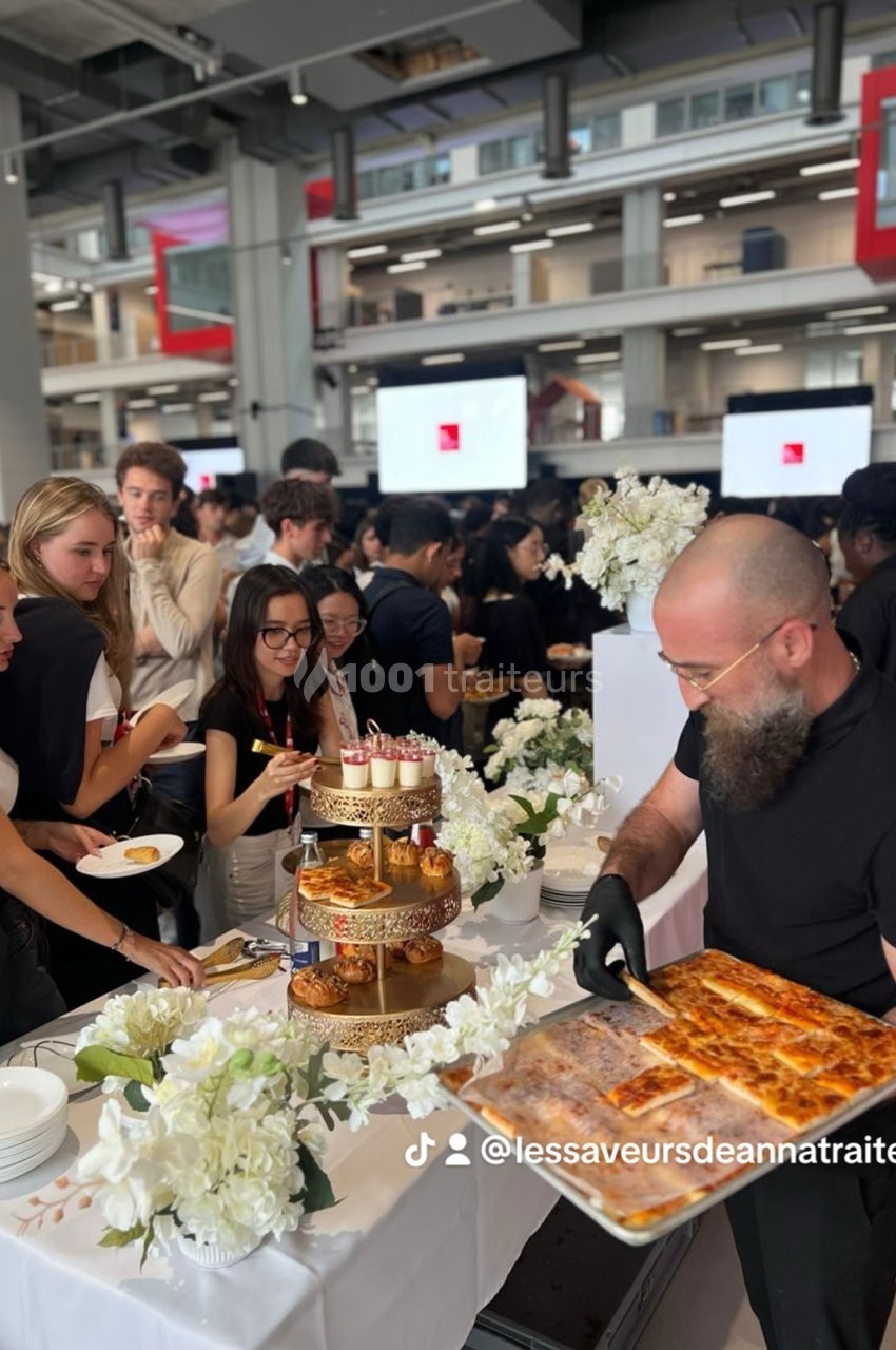 Un traiteur sert des plats lors d'un événement, avec des invités rassemblés autour d'une table décorée de fleurs blanches.