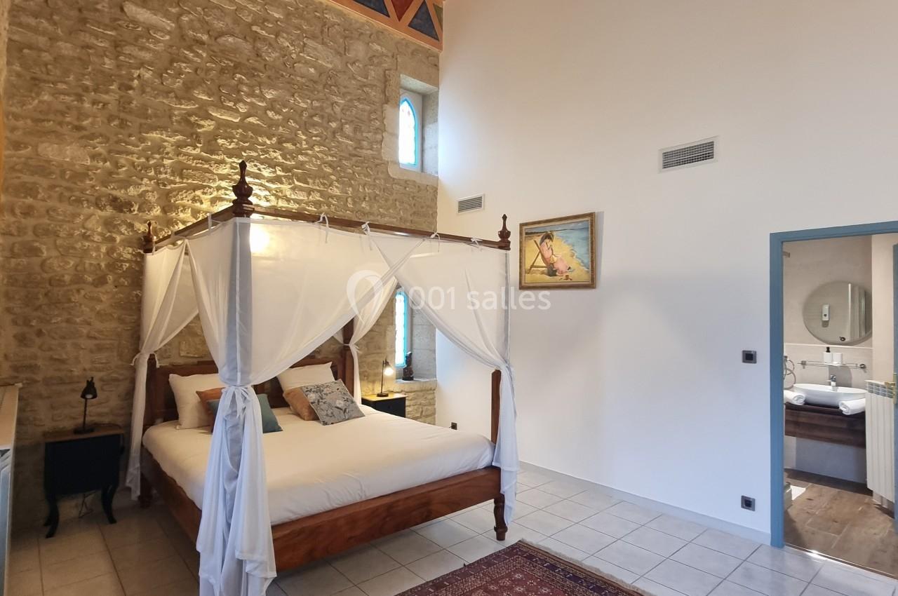 Chambre lumineuse avec lit à baldaquin, murs en pierre apparente, poutres au plafond et tapis au sol.