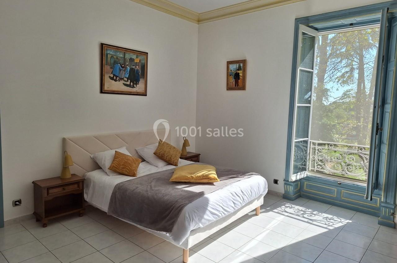 Chambre lumineuse avec un lit double, des coussins jaunes, des tableaux au mur et une fenêtre ouverte sur un balcon.