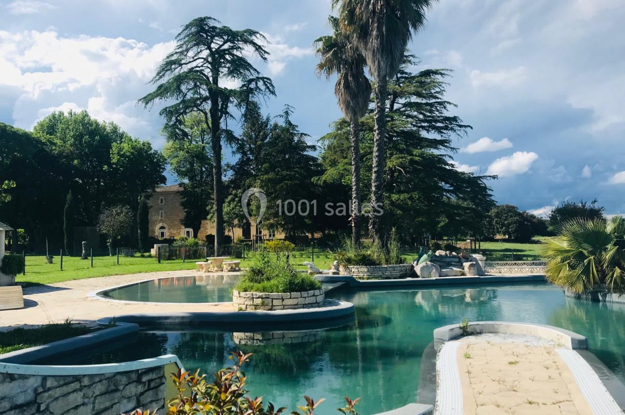 Piscine extérieure entourée de végétation luxuriante, avec des palmiers et un bâtiment en pierre en arrière-plan.