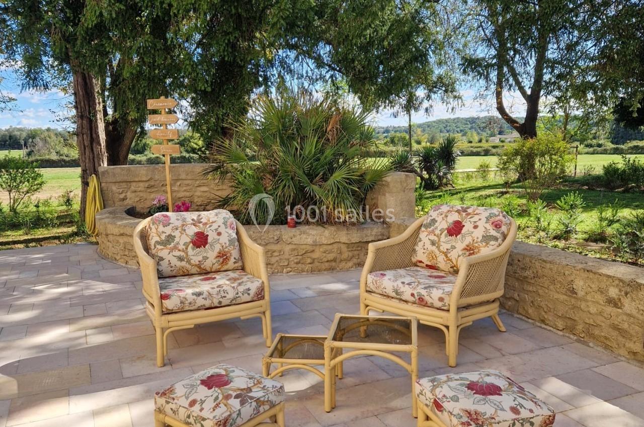 Mobilier en rotin avec coussins fleuris disposé sur une terrasse en pierre, entourée de verdure et d'arbres.