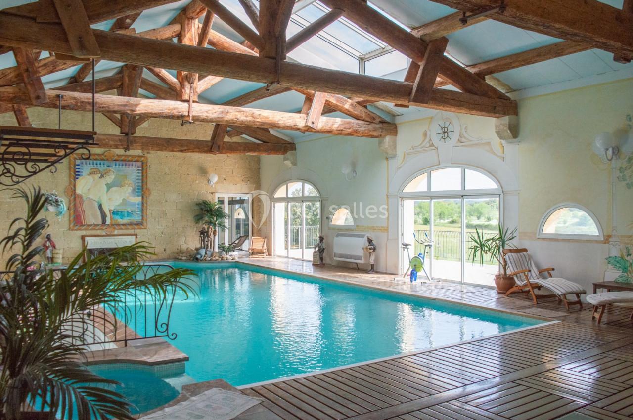 Piscine intérieure lumineuse avec charpente en bois, grandes baies vitrées et chaises longues en bois.