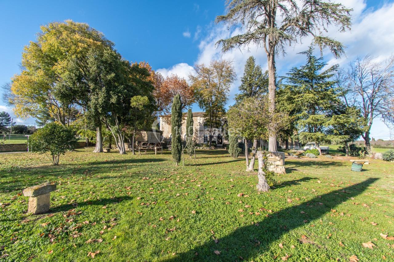 Jardin verdoyant avec arbres variés, pelouse parsemée de feuilles et bâtiment en pierre en arrière-plan.