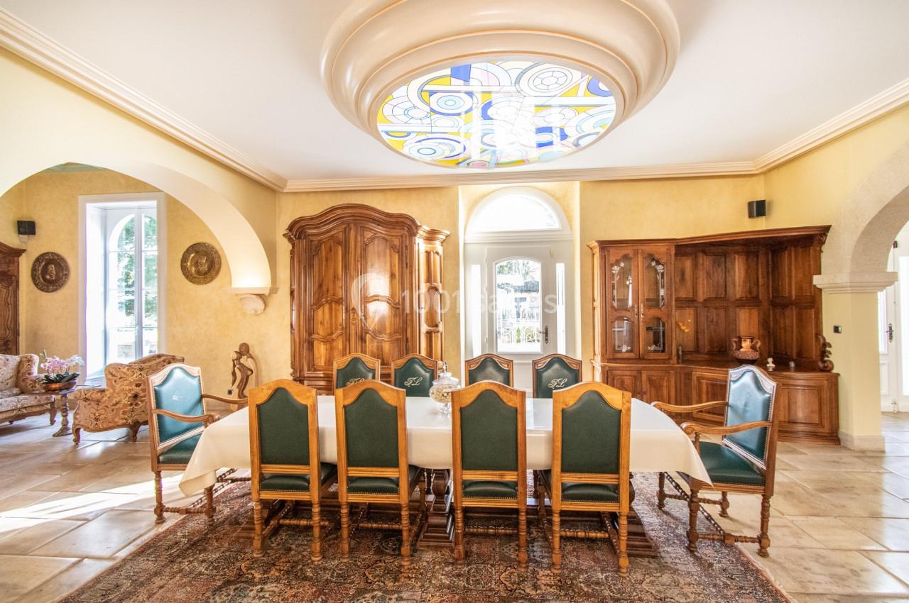 Salle à manger lumineuse avec une grande table entourée de chaises en bois, meubles anciens et plafond décoré d'un vitrail.