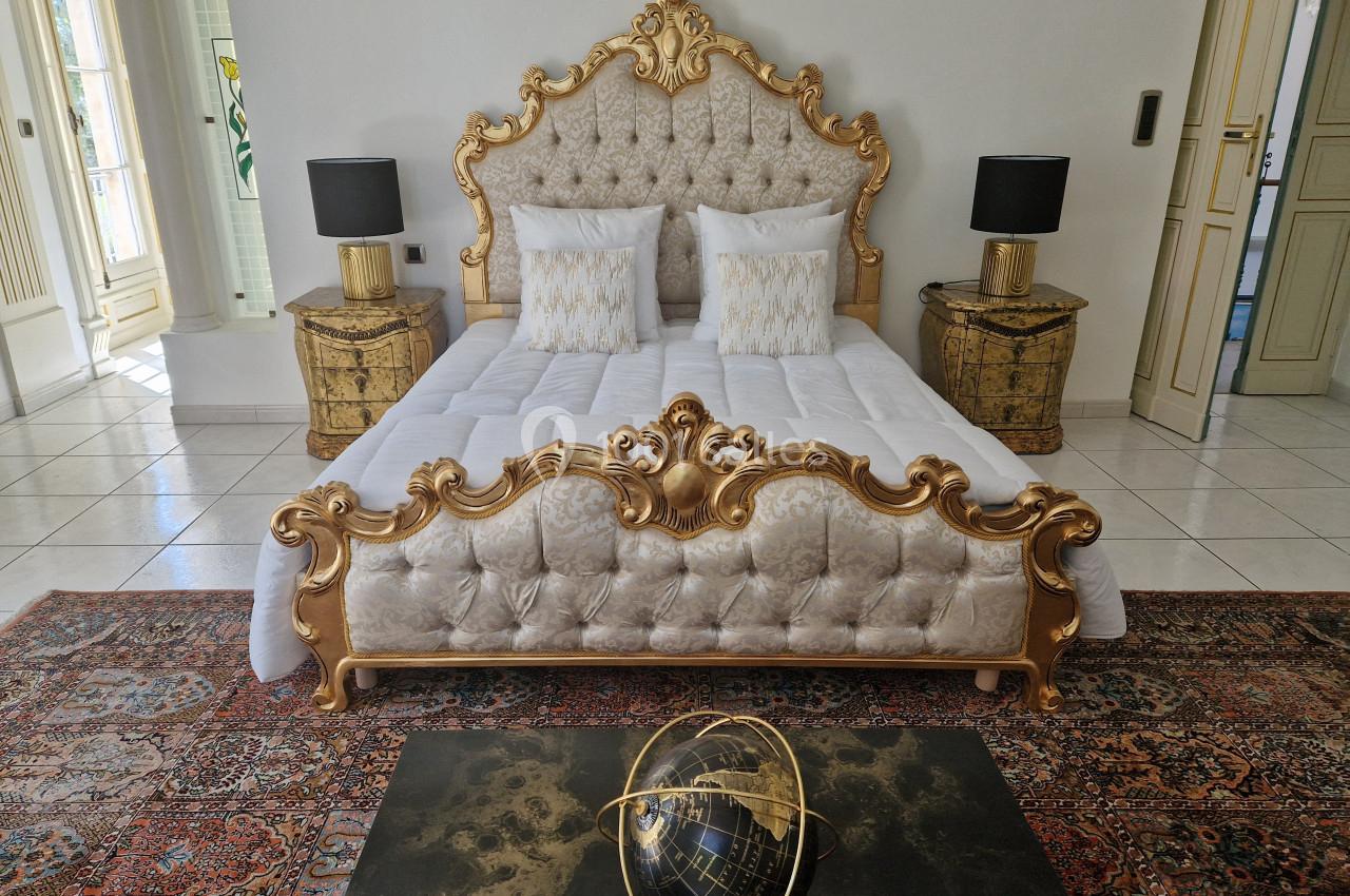 Lit à baldaquin doré avec tête de lit capitonnée, flanqué de tables de chevet et lampes, sur un tapis oriental.