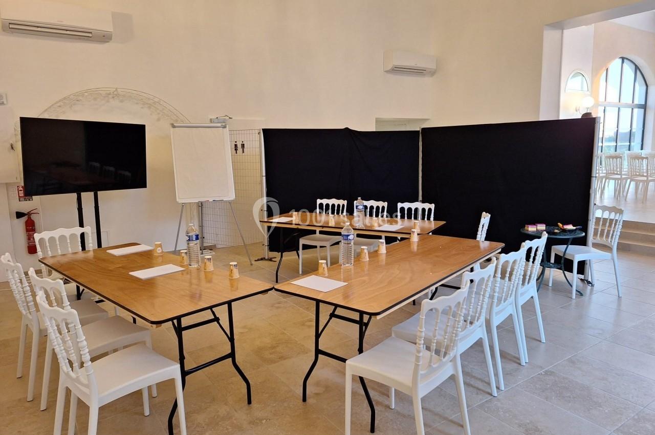 Salle lumineuse aménagée pour une réunion, avec tables en bois, chaises blanches, tableau blanc et écran.