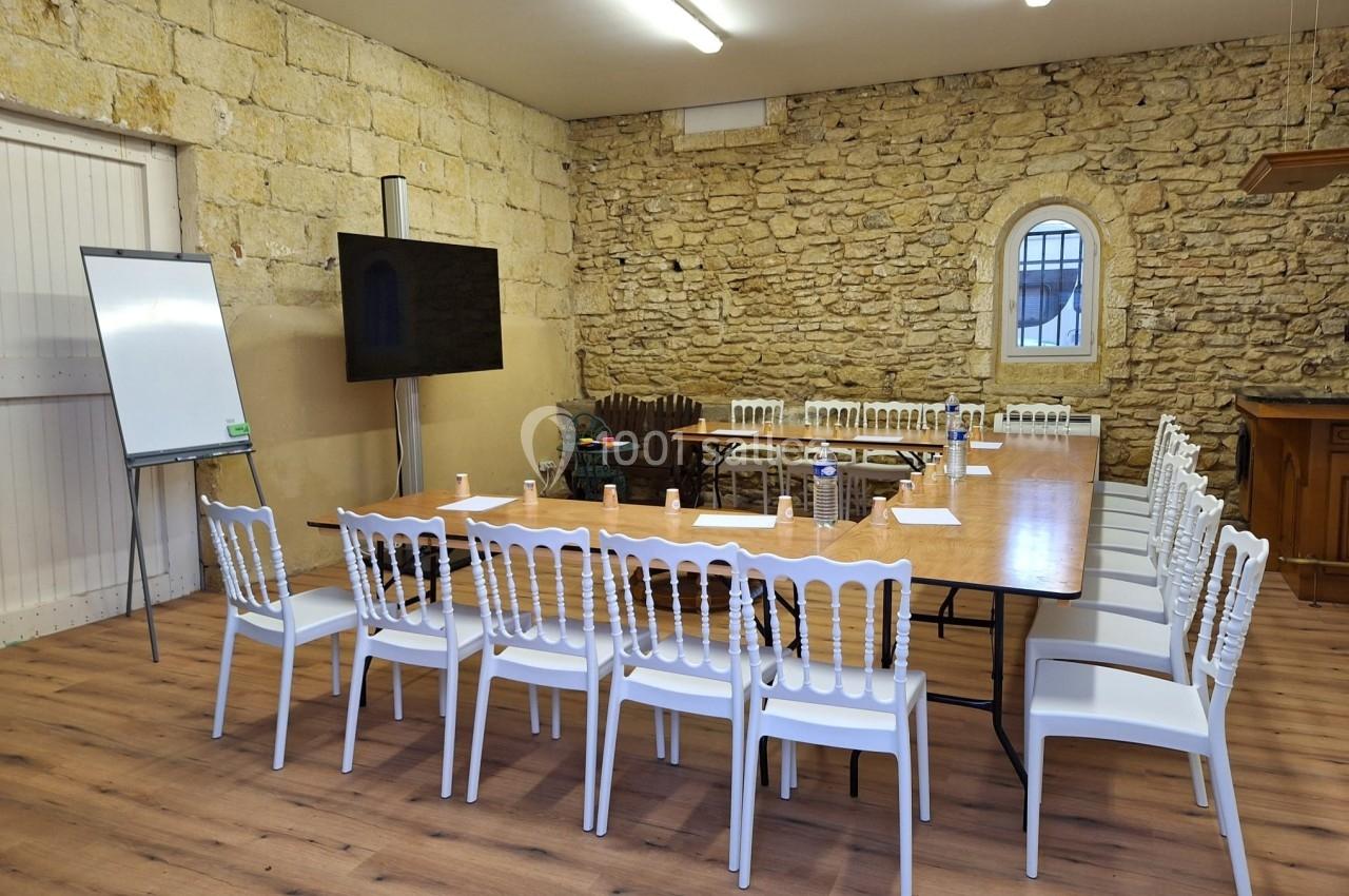 Salle de réunion avec table en bois, chaises blanches, tableau blanc, écran et murs en pierre apparente.