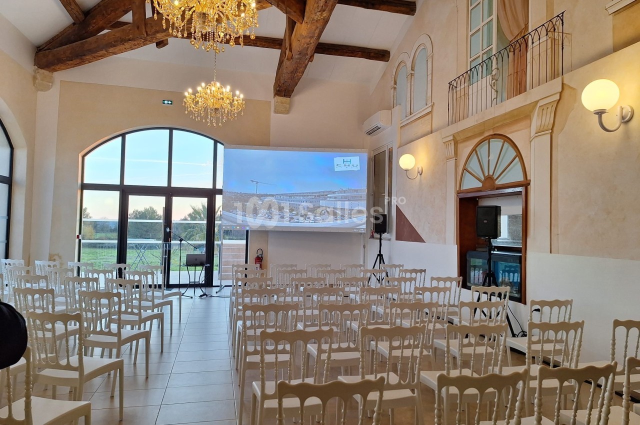 Salle lumineuse avec des chaises blanches alignées, un écran de projection, des lustres et de grandes fenêtres donnant sur l…