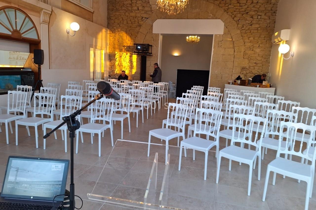 Salle de conférence lumineuse avec des rangées de chaises blanches, un pupitre et un micro au premier plan.