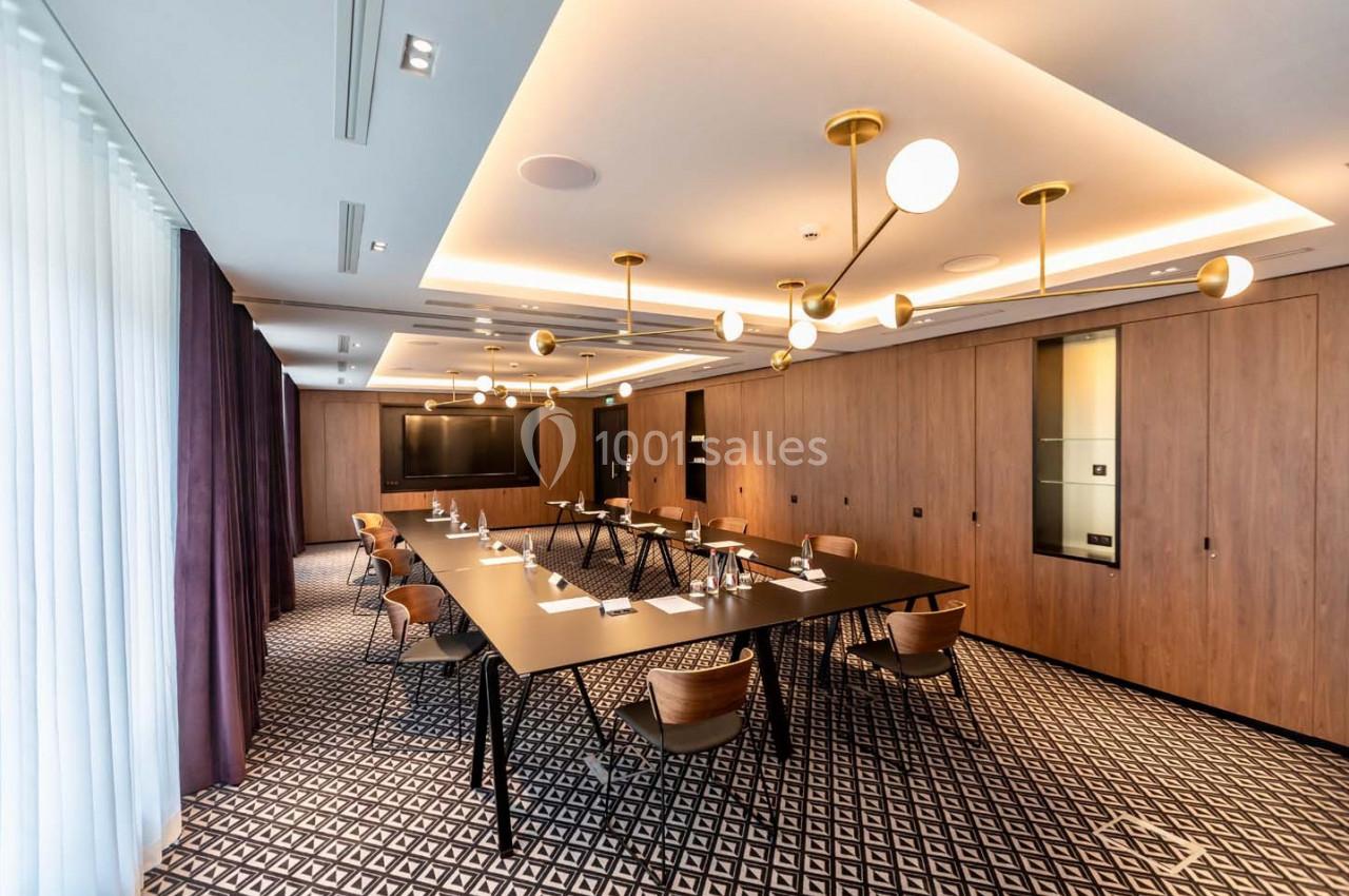 Salle de réunion moderne avec table rectangulaire, chaises en bois, éclairage suspendu et écran mural.