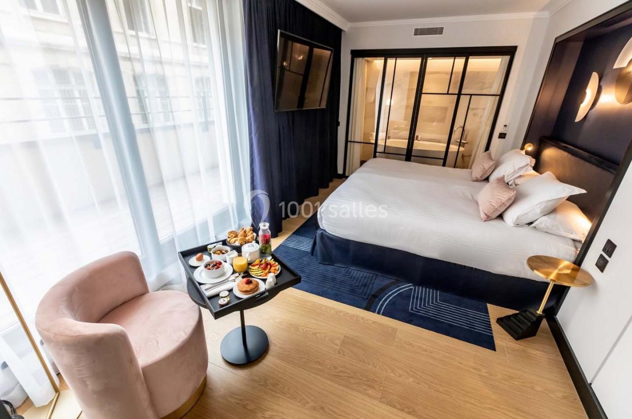 Chambre d'hôtel moderne avec lit double, fauteuil rose, table avec petit-déjeuner et grande baie vitrée.