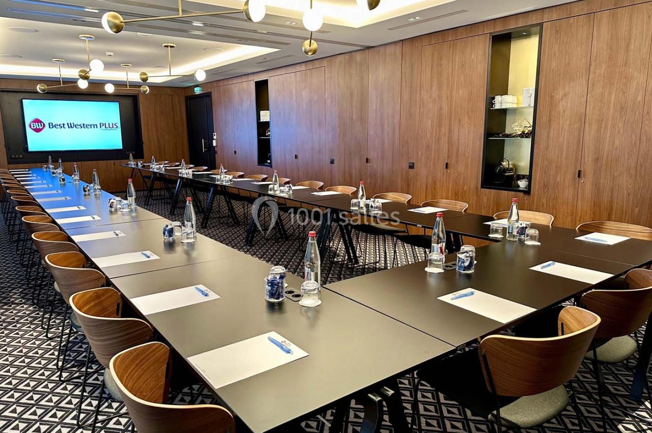 Salle de réunion moderne avec tables disposées en U, chaises en bois, bouteilles d'eau et écran affichant ’Best Western Plus…