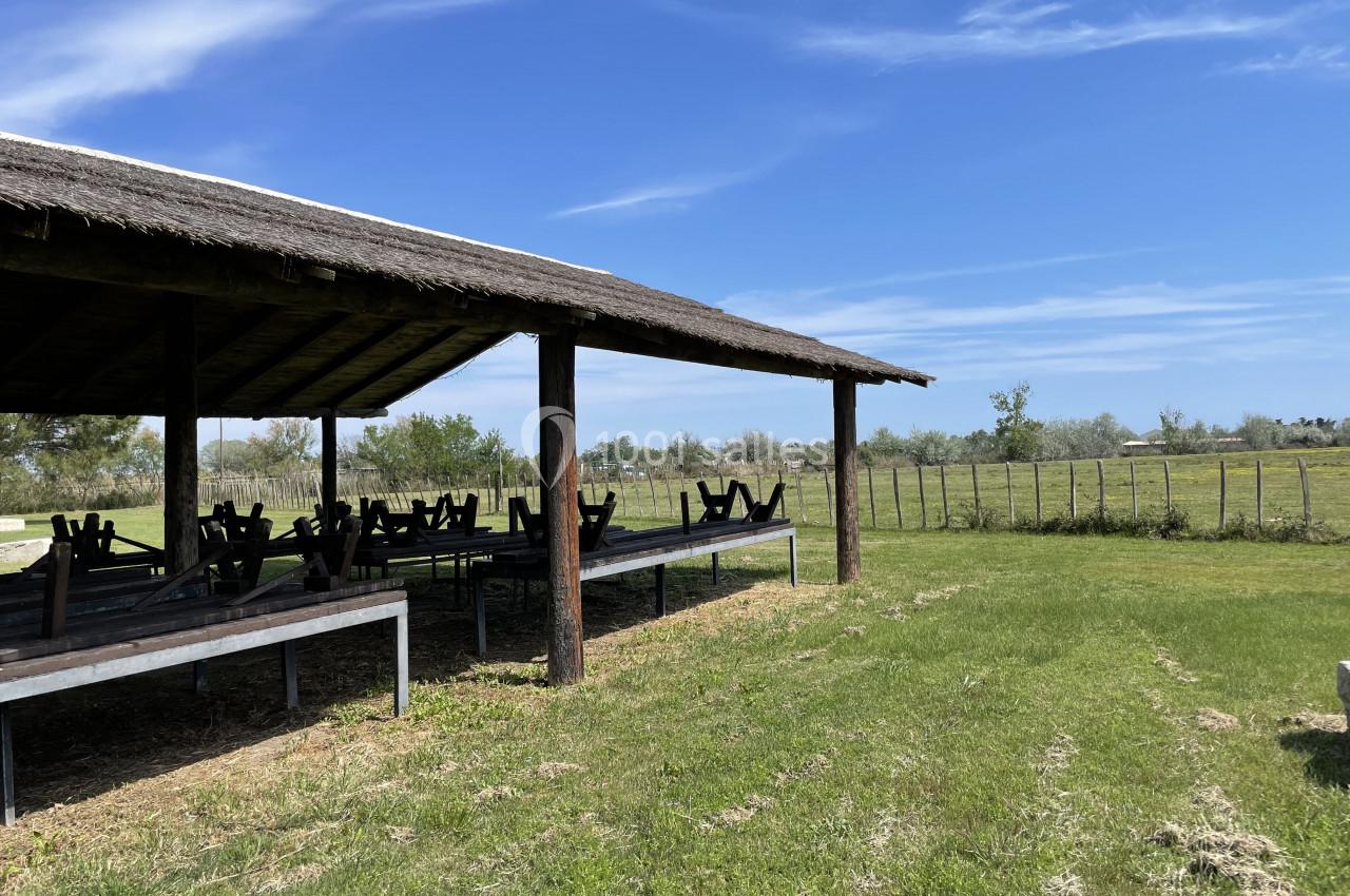 Abri en bois avec tables de pique-nique sous un toit de chaume, entouré d'une prairie et d'une clôture.