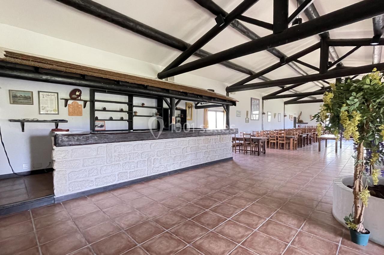 Intérieur d'une salle avec un comptoir en pierre, des poutres apparentes et des tables en bois disposées au fond.