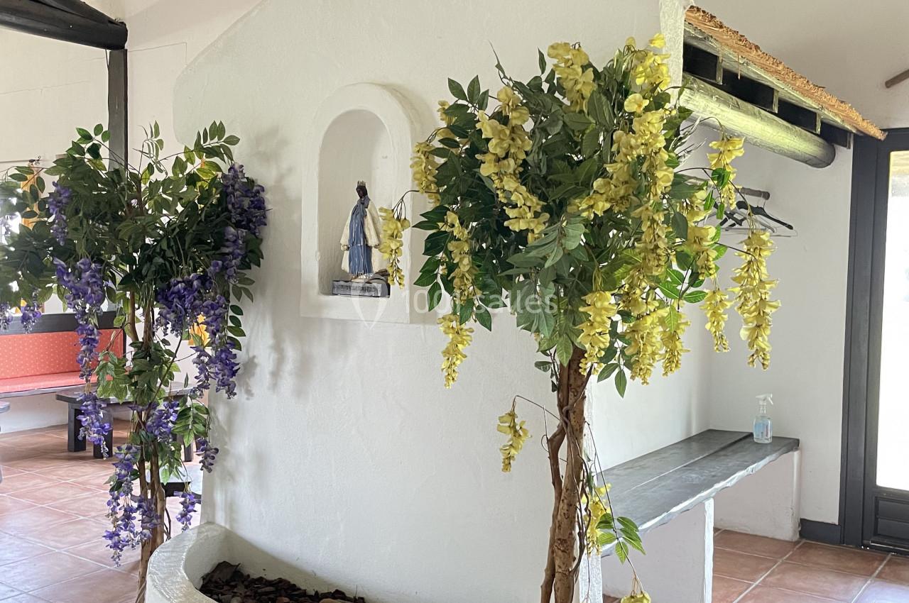 Petite alcôve blanche décorée de plantes artificielles fleuries et abritant une statue religieuse.