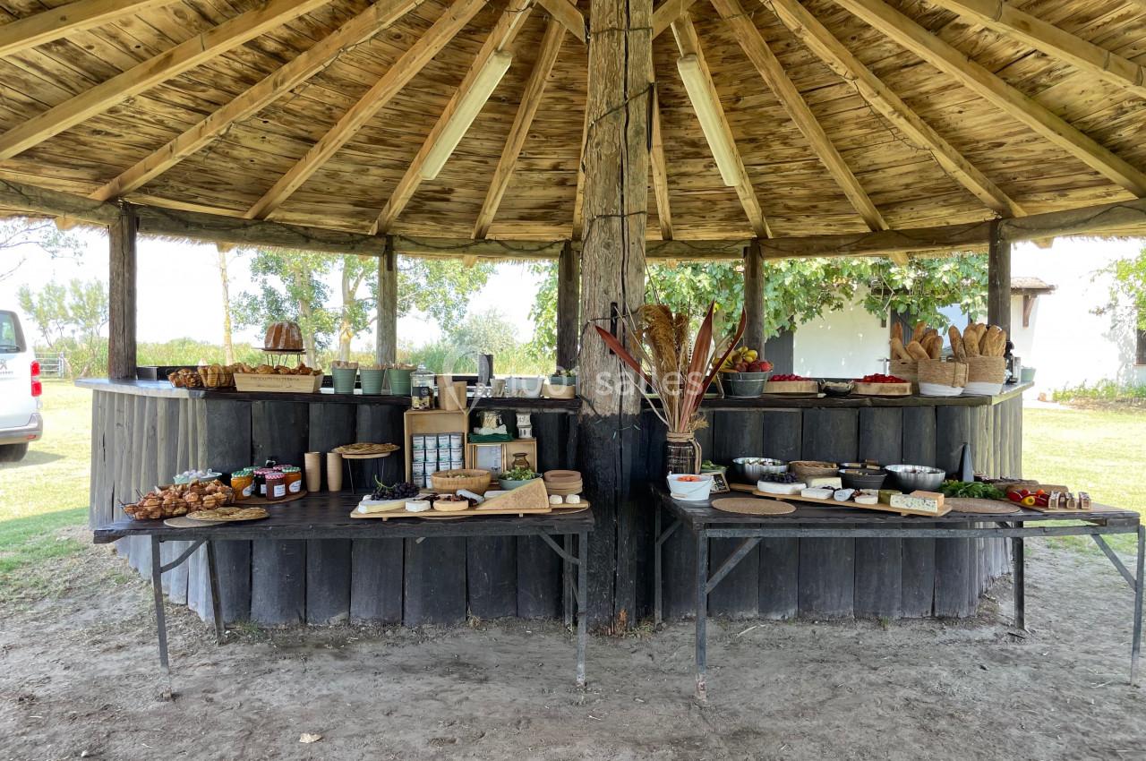 Buffet en plein air sous une structure en bois, proposant pains, confitures, fruits et autres produits alimentaires.