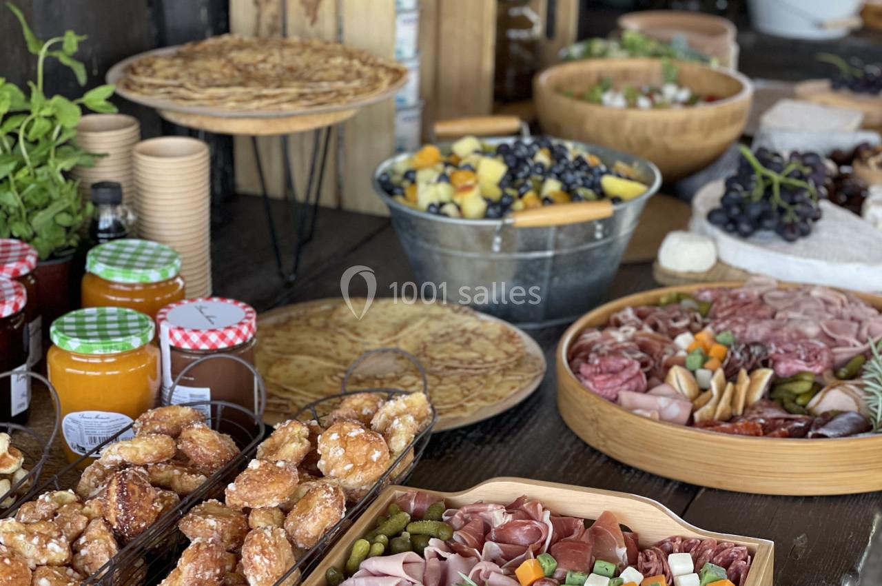 Plateau de charcuterie, fruits, légumes et condiments sur une table en bois avec d'autres plats et boissons en arrière-plan.