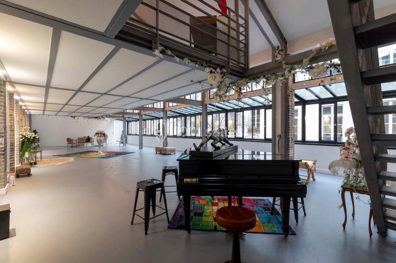 Grand espace lumineux avec verrières, piano noir, tapis coloré et décorations florales, ambiance moderne et élégante.