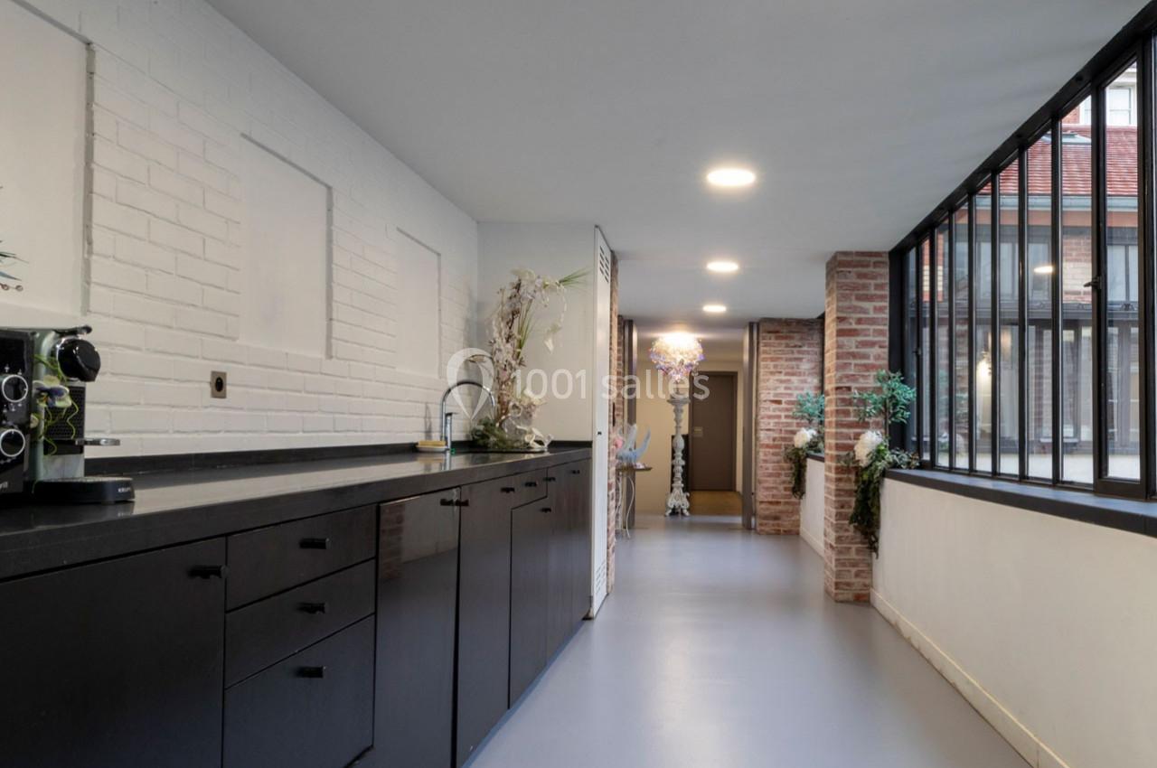 Cuisine moderne avec plan de travail noir, mur en briques blanches, grandes fenêtres et vue sur un couloir lumineux.