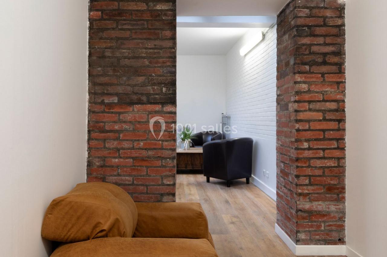 Mur en briques apparentes séparant un espace avec fauteuils marron et noirs sur un sol en bois clair.