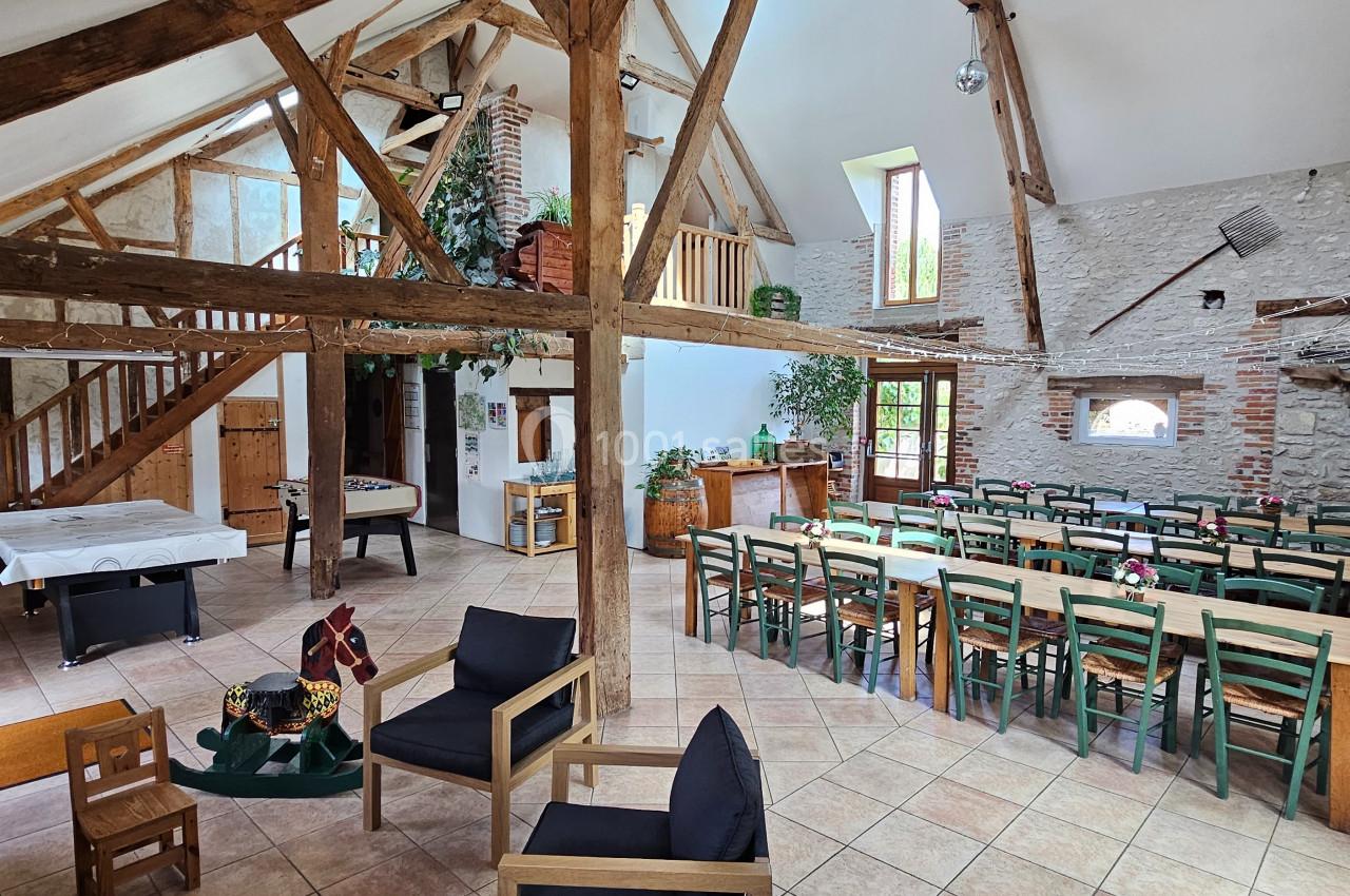 Salle lumineuse avec poutres apparentes, tables en bois, chaises vertes et espace détente dans un cadre rustique.
